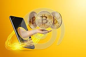 The hand crawling out through the smartphone holds, offers bitcoin coins, yellow background. Crypto currency concept, blockchain