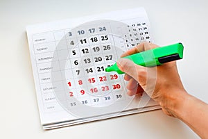 Hand crosses off the green marker days in the calendar. White calendar. Weekends are highlighted in red. Close up photo