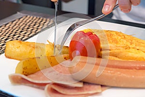 Hand cutting potato on breakfast dish with omelet, sausages, ham, tomato, potatoes fried on white plate