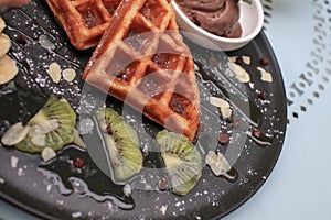 Hand cutting waffle with kiwi and ice cream on black plate. Waffle with honey. Dessert sweetness.