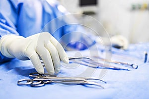 Hand of doctor or surgeon in blue gown pick up surgical clamp instrument or tools inside operating theatre with blur background.