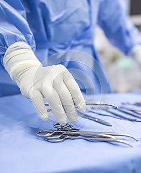 Hand of doctor or surgeon in blue gown pick up surgical clamp instrument or tools inside operating theatre with blur background.