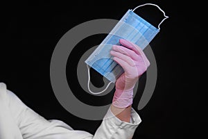 Hand of a doctor in a white coat holding a blue medical mask, close-up on a black background