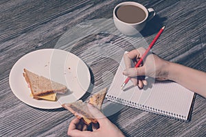 Hand erasing something in a notebook and eat a tasty cheese sandwich. Close up of a plate of sandwich on it and cup of hot cup of