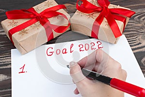 A hand with a felt-tip pen writes a list of New Year`s goals for 2022 on a sheet on a wooden background with boxes of gifts.