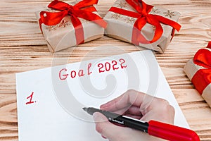 A hand with a felt-tip pen writes a list of New Year`s goals for 2022 on a sheet on a wooden background with boxes of gifts.
