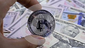 Hand in Fingers Holds a Silver Coin Bitcoin, BTC on a Background with Bills of Dollars