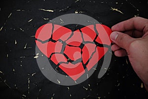 Hand fixing a broken and torn red heart paper cutout in black background.