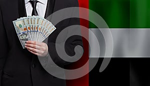 Hand giving 100 US dollar bills money banknotes against flag of United Arab Emirates background