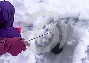 The hand of a gloved angler girl in winter holds a small fishing rod with a nod to a hole in the ice.