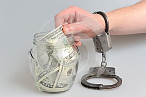 Hand in handcuffs taking money from glass jar on gray