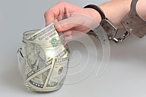 Hand in handcuffs taking money from glass jar on gray