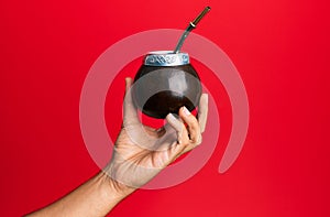 Hand of hispanic man holding cup of mate infusion beverage over isolated red background