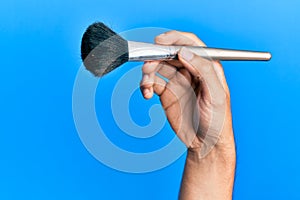 Hand of hispanic man holding makeup brush over isolated blue background