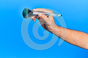 Hand of hispanic man holding makeup brush over isolated blue background