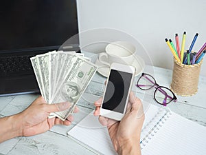 Hand hold money dollar and mobilephone with workspace on white wood table background