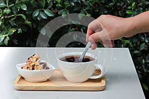Hand hold spoon stirring coffee on wood tray and there is cookie at side. Table is set in garden with green leaves background.