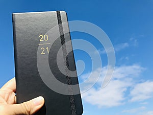 A Hand holding a black 2021 diary book with blue sky background.