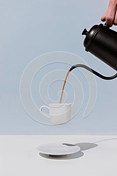 A hand holding black metal pot and pouring coffee into levitating white cup