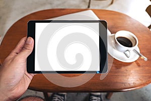 Hand holding black tablet pc with blank desktop white screen with laptop and coffee cup on wooden table