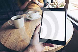 A hand holding black tablet pc with white blank screen on wooden table with woman using mobile phone in backgroun