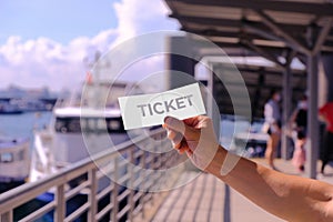 Hand holding blank mockup for one-fold ticket design, at a pier. Cruise ferry and incidental people in the background, defocused.