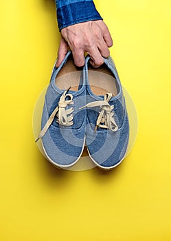 Hand holding blue sneakers isolated on a yellow background. Sports shoes hanging.