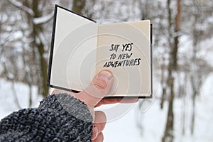 Hand holding a book with the inscription Say yes to new adventures on the background of the winter forest