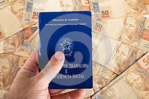 Hand holding Brazilian document work and social security Carteira de Trabalho e Previdencia Social with fifty reais banknotes.