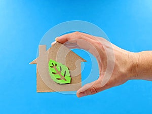 Hand holding a cardboard house with green leaf inside on blue background, concept image of energy-efficient