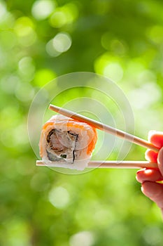 Hand holding chopsticks sushi roll with red fish on summer natural background