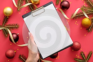 Hand holding clipboard mockup with blank paper over Christmas background with Christmas decorations, balls, gold ribbon, fir tree