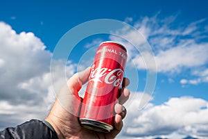 Hand holding a Coca Cola can towards bright blue and cloudy sky outdoors