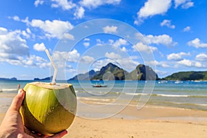 Hand holding a coconut coco loco coctail with straw, tropical islands, beach, sea and blue sky in the background, El Nido, Palawan