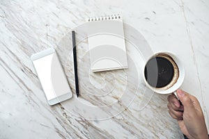 A hand holding coffee cup with a white mobile phone with blank desktop screen and empty notebook on table