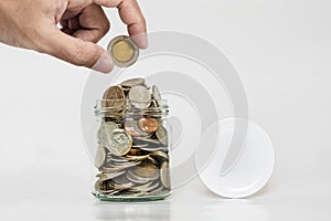 Hand holding coin with jar glass full of coins, and jar cap with copy space