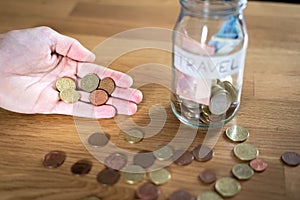 Hand holding coins and a jar next to it labeled travel with euro money inside and coins outside on top of a wooden table.