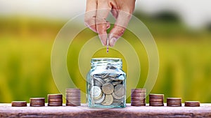 Hand holding coins in a jar for saving money and stacks of coins on the wooden floor