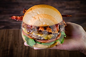 Hand holding a craft beef burger with cheese, bacon, caramelized onion and rocket leafs on rustic background