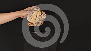 The hand is holding a crumpled brown paper maul and trash can on black background