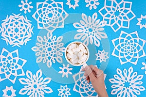Hand holding cup of hot chocolate with marshmallow and paper snowflakes on blue background. Top view. Christmas decoration