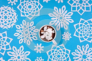 Hand holding cup of hot chocolate with marshmallow and paper snowflakes on blue background. Top view. Christmas decoration