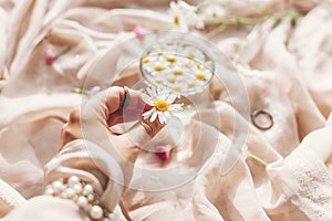 Hand holding daisy flower on background of soft beige fabric with glass cup with flowers and jewelry in sunny light. Tender floral