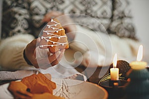 Hand holding decorated christmas tree gingerbread cookie on background of rustic table with napkin, candle, decorations. Moody