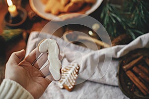 Hand holding decorated gingerbread cookie candy cane on background of rustic table with napkin, candle, decorations. Moody image.