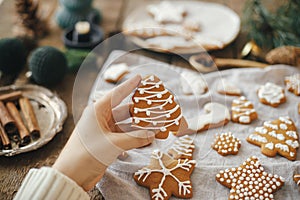 Hand holding decorated gingerbread cookie christmas tree on background of rustic table with napkin, candle, decorations. Moody