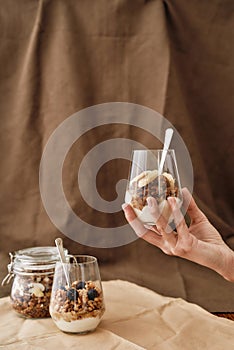 Hand holding delicious layered dessert in glass jar, Homemade yogurt with granola and berries, Granola baked with nuts