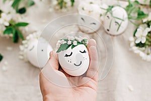 Hand holding easter egg with drawn sleeping face in floral wreath on soft rustic background