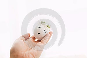 Hand holding easter egg with drawn sleeping face in floral wreath on white background in soft light