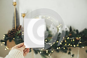 Hand holding empty greeting card on background of stylish christmas houses, fir branches with golden lights and tree decorations